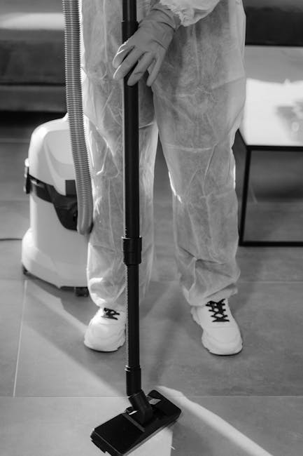 A person wearing protective gloves and disposable coveralls is performing surface cleaning in an indoor space, using a vacuum cleaner with a floor attachment. The area features a smooth, polished floo