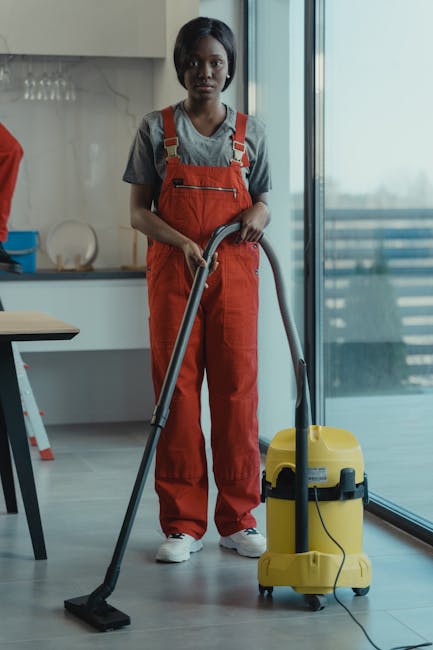 A person wearing protective gloves and disposable coveralls is performing surface cleaning in an indoor space, using a vacuum cleaner with a floor attachment. The area features a smooth, polished floo