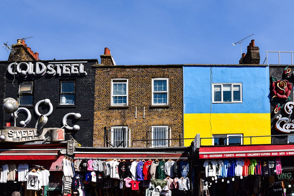A colorful street scene featuring a row of shops with varied facades, including a black building with the word 'COLDSTEEL' displayed in white 3D letters, adjacent to a brown brick building and a vibra